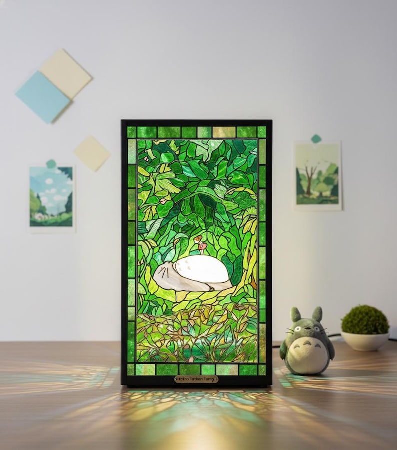 May include: A stained glass lamp featuring a green forest scene with a white creature resting. The lamp has a black frame and the text "Retro Tatten Tang" at the bottom. A small Totoro figure and a small plant are next to the lamp.