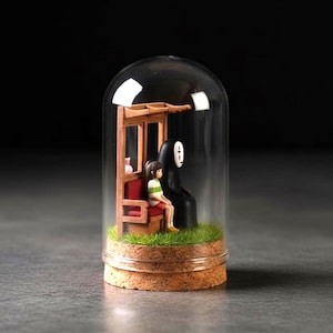 Puede incluir: Un diorama en miniatura dentro de una c&uacute;pula de cristal con personajes de una popular pel&iacute;cula de animaci&oacute;n. La escena incluye una ni&ntilde;a, un esp&iacute;ritu sin rostro y una estructura de parada de autob&uacute;s, todo sobre un lecho de hierba verde. La base es de corcho.