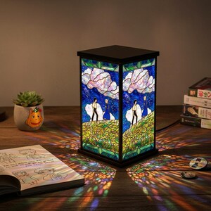May include: A stained glass lamp with a black frame. The lamp features a scene of two figures on a hill under a starry sky with large white clouds. The lamp is on a wooden surface, casting colorful light patterns.