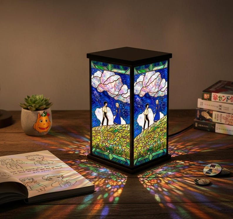 May include: A stained glass lamp with a black frame. The lamp features a scene of two figures on a hill under a starry sky with large white clouds. The lamp is on a wooden surface, casting colorful light patterns.