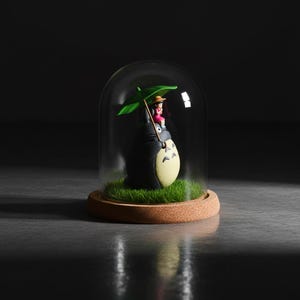 Puede incluir: Una c&uacute;pula de cristal decorativa con una figura de Totoro y una ni&ntilde;a sosteniendo un paraguas verde. La figura est&aacute; sobre c&eacute;sped artificial, sobre una base de corcho. La c&uacute;pula es transparente y el fondo es oscuro.