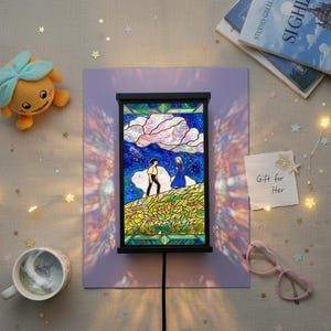 May include: A stained glass lamp with a black frame, depicting a couple on a hill under a starry sky. The lamp is on a purple background, with a plush toy, a mug, and a note that says "Gift for Her".