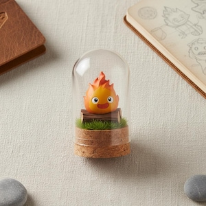 Puede incluir: Una campana de cristal decorativa con un personaje de dibujos animados con llamas naranjas, una cara sonriente y una base de tronco. La campana se asienta sobre una base de corcho con c&eacute;sped artificial. Un cuaderno encuadernado en cuero y una peque&ntilde;a piedra est&aacute;n en el fondo.