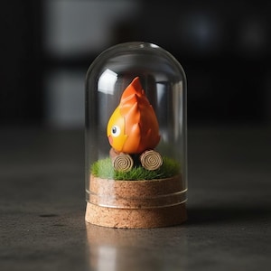 Puede incluir: Una caprichosa figura de personaje naranja y amarillo con una cabeza en forma de llama, dentro de una c&uacute;pula de cristal. La figura descansa sobre un lecho de musgo verde y dos troncos de madera, sobre una base de corcho.