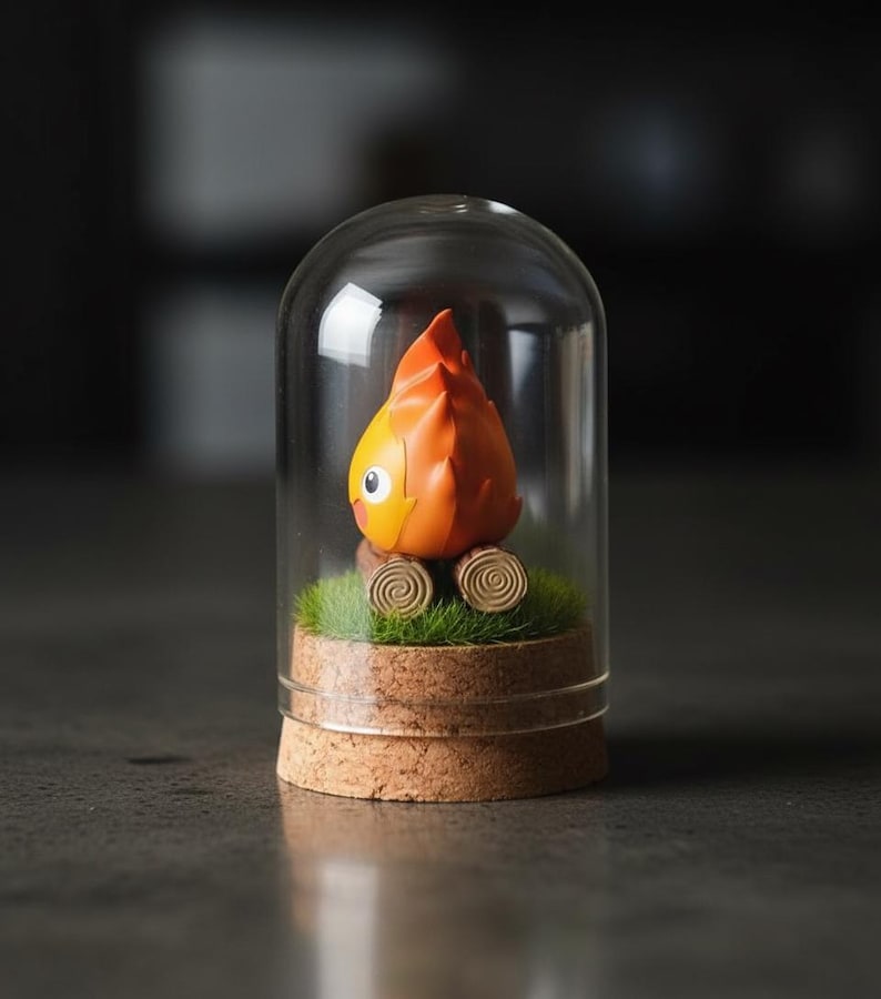 Puede incluir: Una caprichosa figura de personaje naranja y amarillo con una cabeza en forma de llama, dentro de una c&uacute;pula de cristal. La figura descansa sobre un lecho de musgo verde y dos troncos de madera, sobre una base de corcho.