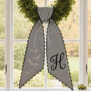 May include: A green boxwood wreath adorned with a black and white striped fabric ribbon tied in a knot. The ribbon features black lace trim, a black embroidered letter H on one side, and four small gray bat silhouettes on the other side.