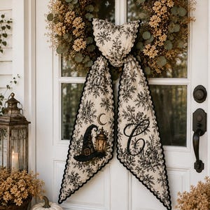 May include: A fabric door hanger featuring a cream and black toile pattern with a witch hat, a lantern, a crescent moon, and a large letter C. The piece has a scalloped black edge and is draped over a large, round, dried floral wreath.