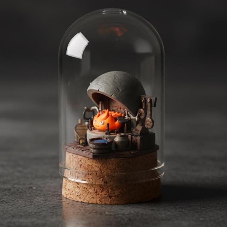 Puede incluir: Diorama en miniatura de una criatura de fuego dentro de una c&uacute;pula de cristal. La escena incluye una forja en miniatura, herramientas y una peque&ntilde;a olla azul. La base es de corcho. La c&uacute;pula es transparente y el fondo es oscuro.