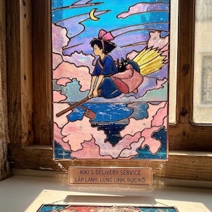 May include: Stained glass panel featuring a young girl on a broomstick, flying through a night sky with a crescent moon. The panel has a pink, blue, and purple color scheme, with the text "Kiki's Delivery Service" at the bottom.