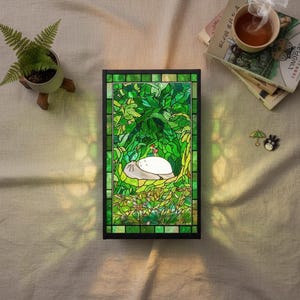 May include: A stained glass panel with a black frame, depicting a scene with a large, white creature resting in a lush green forest. The panel is illuminated, casting a warm glow. A small potted fern and a cup of tea are nearby.