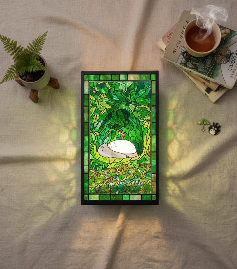 May include: A stained glass panel with a black frame, depicting a scene with a large, white creature resting in a lush green forest. The panel is illuminated, casting a warm glow. A small potted fern and a cup of tea are nearby.