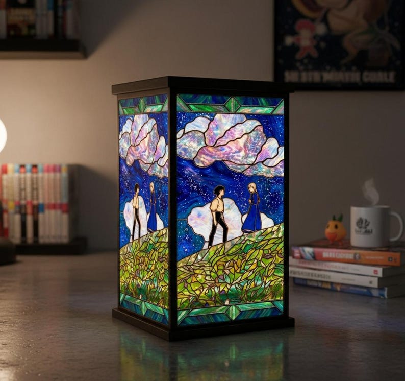 May include: A rectangular stained glass lamp with a black frame. The lamp features a colorful scene of two figures on a hill under a starry sky with fluffy clouds. The lamp is on a table.