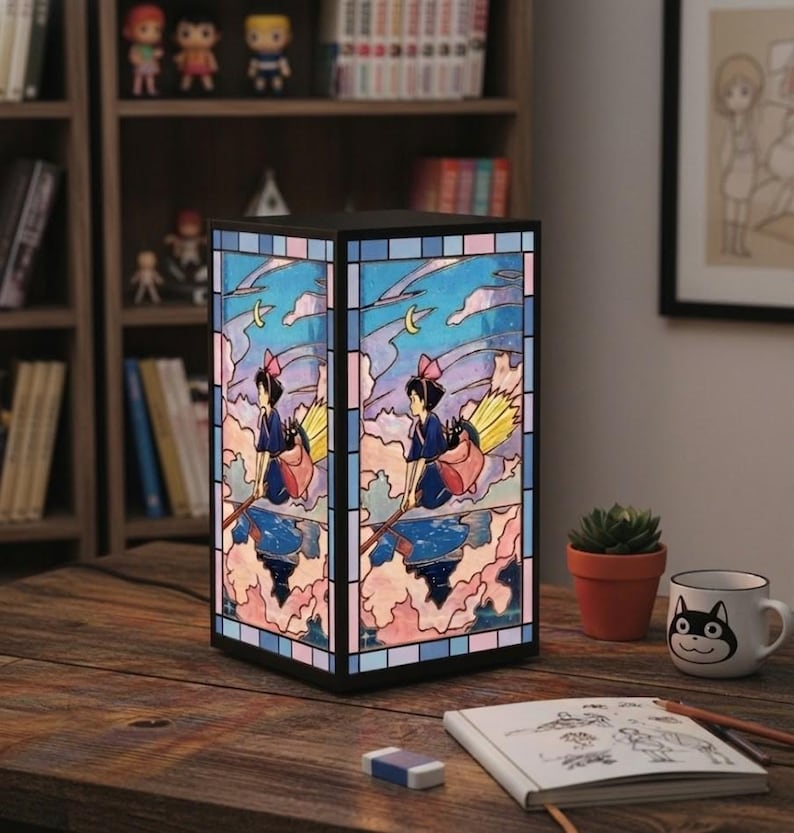 May include: A stained glass lamp featuring a cartoon witch on a broomstick against a cloudy sky background. The lamp has a black frame and is placed on a wooden table. A small succulent and a cat-themed mug are also on the table.