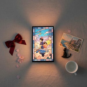 May include: A framed stained-glass panel depicts a young witch flying on a broom over clouds, with a crescent moon. A red bow, pink flower petals, a postcard, a black cat figurine, and a white teacup are arranged around the panel.