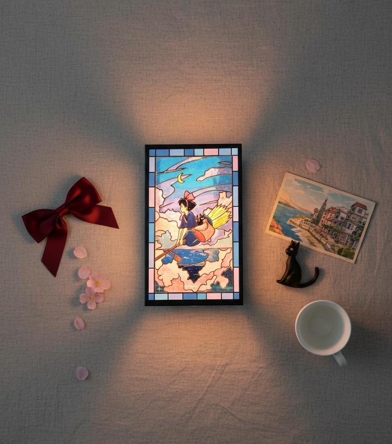 May include: A framed stained-glass panel depicts a young witch flying on a broom over clouds, with a crescent moon. A red bow, pink flower petals, a postcard, a black cat figurine, and a white teacup are arranged around the panel.