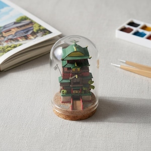 May include: A miniature, multi-tiered building model with a green and red color scheme, encased in a glass dome. The model sits on a cork base. A book and paint palette are in the background.