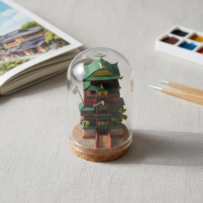 May include: A miniature, multi-tiered building model with a green and red color scheme, encased in a glass dome. The model sits on a cork base. A book and paint palette are in the background.