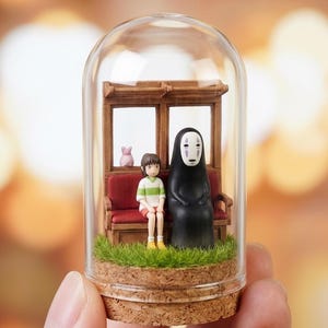 Puede incluir: Una c&uacute;pula de cristal en miniatura con personajes de una pel&iacute;cula de animaci&oacute;n popular. En el interior, una ni&ntilde;a con una camisa a rayas verdes y blancas est&aacute; sentada junto a una figura negra. Un cerdo rosa y un banco de madera tambi&eacute;n est&aacute;n presentes.