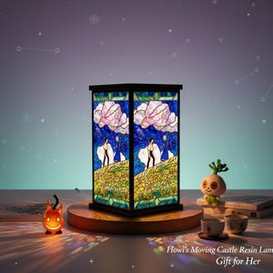 May include: A square resin lamp with a stained-glass design featuring characters and scenery from "Howl's Moving Castle." The lamp has a black frame and sits on a wooden base. A small pumpkin-shaped light and other decorative items are nearby.