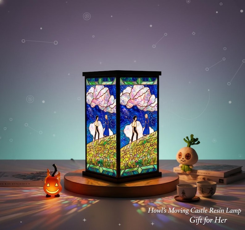 May include: A square resin lamp with a stained-glass design featuring characters and scenery from "Howl's Moving Castle." The lamp has a black frame and sits on a wooden base. A small pumpkin-shaped light and other decorative items are nearby.