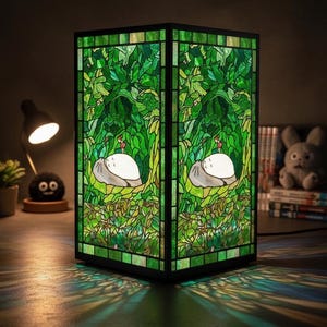 May include: A stained glass lamp with a black frame. The lamp features a green and white design with a forest scene and a white creature. The lamp is illuminated, casting green and blue light patterns on the surface below.