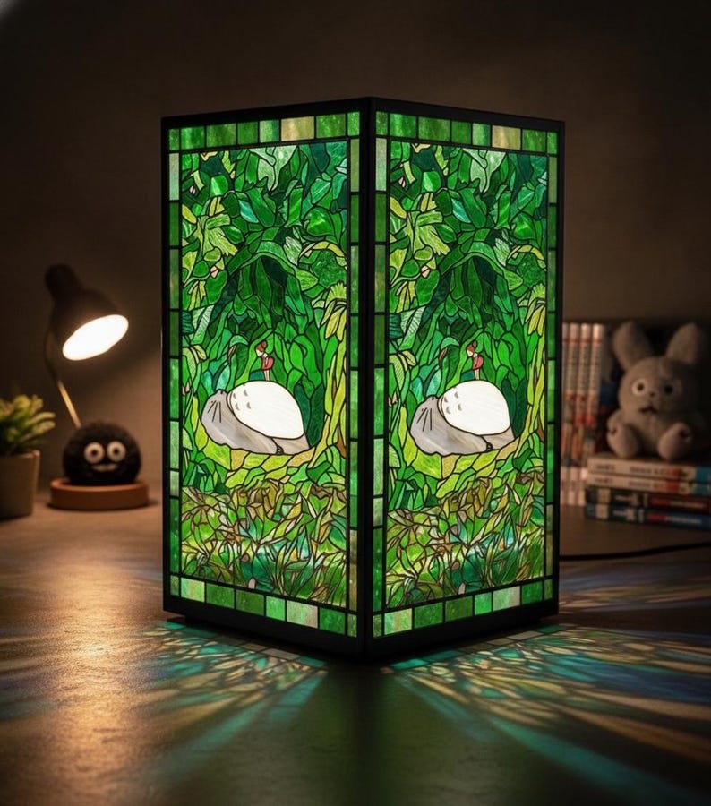 May include: A stained glass lamp with a black frame. The lamp features a green and white design with a forest scene and a white creature. The lamp is illuminated, casting green and blue light patterns on the surface below.