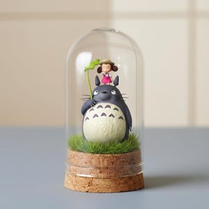 Puede incluir: Una c&uacute;pula de cristal decorativa con una figura de Totoro y una ni&ntilde;a peque&ntilde;a sosteniendo una hoja. Las figuras est&aacute;n sobre un lecho de c&eacute;sped artificial verde, sobre una base de corcho. La c&uacute;pula es transparente y las figuras est&aacute;n detalladas.