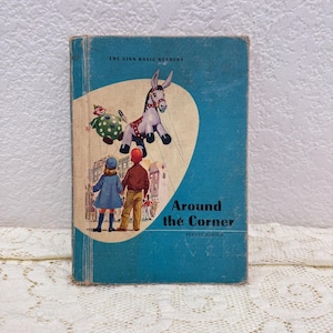Könnte beinhalten: Vintage Kinderbuch mit dem Titel "Around the Corner" mit blauem Einband. Das Cover zeigt Illustrationen eines Marionetten-Esels, eines Clowns und zweier Kinder. Das Buch ist eine "Revised Edition" aus der Reihe "The Ginn Basic Readers".