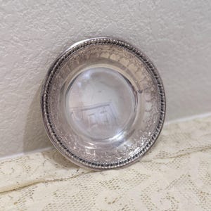 May include: A circular silver-toned metal dish featuring a smooth, reflective center basin and a wide, decorative rim. The outer edge is finished with a textured rope-style border, while the inner rim displays an intricate, embossed floral pattern.