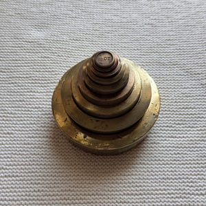 May include: A set of nested brass weights stacked in a conical shape. The top weight is marked with the text 20 oz and a small symbol. The metal has a weathered, golden-brown patina with visible surface scratches and signs of age and use.