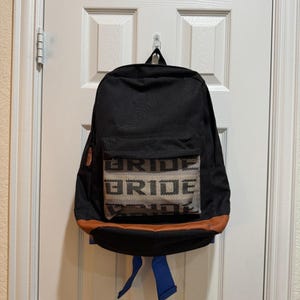 Pode incluir: Mochila preta com um bolso frontal com a palavra "BRIDE" em um padrão repetido. A mochila tem acabamento laranja e alças azuis, pendurada em uma porta branca.