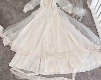 Puede incluir: Un vestido blanco largo hasta el suelo con mangas largas transparentes, cuello alto con detalles de encaje intrincados y una falda de varias capas con ribete de encaje. Un velo blanco transparente a juego con un detalle floral se sitúa al lado.