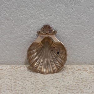 Peut inclure: Un plat en forme de coquillage doré métallique avec un bord festonné et une crête décorative en éventail sur le dessus. L'intérieur du plat présente des rainures verticales profondes rayonnant depuis la base, avec une finition polie.