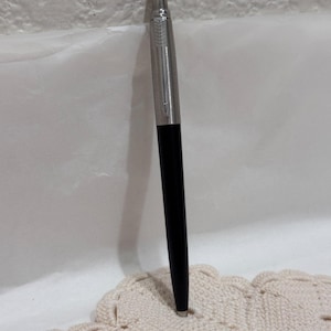 Könnte beinhalten: Ein schwarzer und silberner Stift mit silberner Kappe. Der Stift hat ein schlankes Design und ist vor einem weißen Hintergrund platziert. Ein klassisches Schreibgerät.