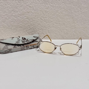 Puede incluir: Gafas con montura dorada y lentes ovaladas, junto a un estuche azul claro con un estampado floral y de mariposas. El estuche rectangular tiene una parte superior curvada. Las gafas están sobre una superficie blanca.