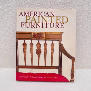 Pode incluir: Um livro de capa dura intitulado American Painted Furniture de Cynthia V. A. Schaffner e Susan Klein. A capa em tom off-white apresenta uma ilustração castanha de um banco de madeira com detalhes florais e o título em letras douradas.