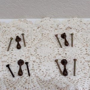 Puede incluir: Cuatro juegos de tiradores de cajón antiguos de bronce con tornillos a juego. Cada tirador tiene un diseño decorativo y forma de lágrima. Los tornillos son una mezcla de latón y metal oscuro. Dispuestos sobre un mantel de encaje blanco.