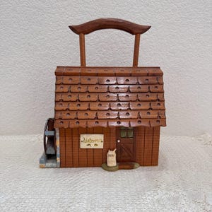 May include: A wooden miniature watermill with a brown shingled roof, a water wheel, and a sign that reads "Watermill". A small bag labeled "MEAL" sits near the door. The handle is arched.