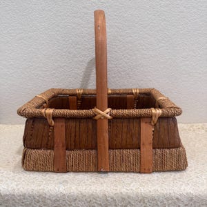 May include: A rectangular basket featuring a tall, centered wooden handle. The body is constructed from dark brown woven fibers with a lighter, rope-like texture along the base. Vertical wooden slats reinforce the sides and connect to the handle.