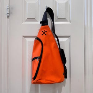 May include: Bright orange sling bag with a black strap and trim. The bag has a zippered front pocket and a small logo near the top. The bag is hanging on a white door.