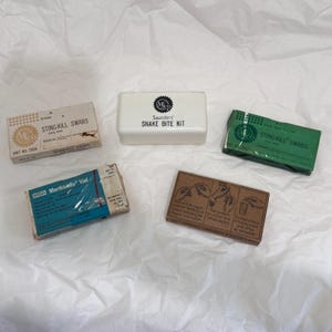 Op de afbeelding: Een verzameling van vijf vintage medische voorraaddoosjes, waaronder een witte Saunders slangenbeetset, twee Sting-Kill wattenstaafjesdoosjes in beige en groen, een blauw MSA Merthiolate flacon-doosje en een bruin doosje met geïllustreerde gebruiksinstructies.