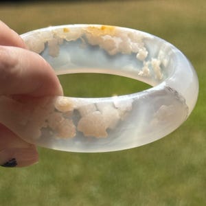 Puede incluir: Un brazalete rígido de color blanco translúcido con inclusiones internas nubladas y sutiles manchas de color ámbar. La pieza de joyería circular tiene una superficie lisa y pulida con un perfil redondeado, mostrando patrones minerales naturales.