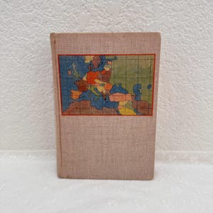 May include: A hardcover book with a light beige fabric cover featuring a rectangular inset map of Europe. The map displays various countries in muted shades of blue, green, orange, and yellow, overlaid with a thin dark grid pattern.