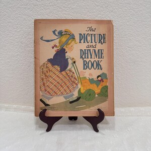 May include: A vintage book titled The Picture and Rhyme Book with a tan cover featuring an illustration of a blonde child in a blue coat and plaid skirt pushing a yellow toy carriage containing two smaller figures with green wheels.