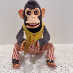 Op de afbeelding: Vintage opwindbare speelgoed aap met een gele vest, rood-wit gestreepte broek en bekkens. De aap heeft een zwarte kop, grijze armen en een lachend gezicht. Een klassiek verzamelobject.