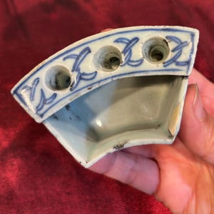 May include: A small, antique, blue and white porcelain inkwell with three holes. The inkwell has a curved shape and is decorated with blue floral patterns. The inkwell is held in a hand.