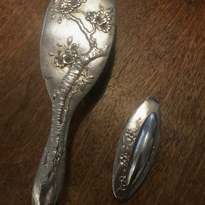 May include: Silver-toned vanity set featuring a hairbrush and a small hand mirror. Both pieces are adorned with an embossed floral and tree branch design. The brush has a long handle, while the mirror is oval-shaped.
