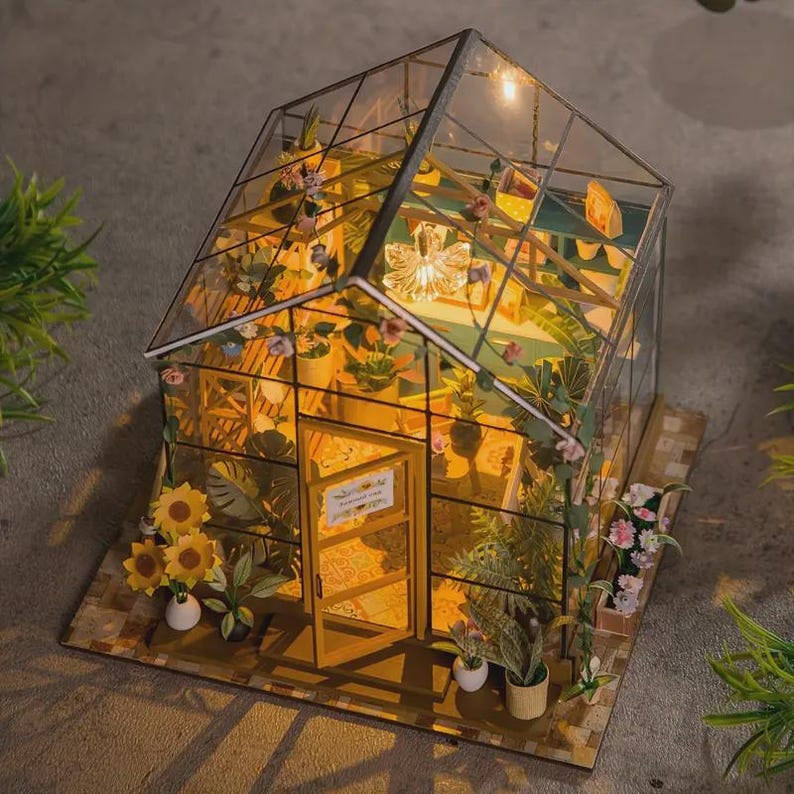 May include: Miniature greenhouse model with glass walls and a wooden frame. The interior is filled with tiny plants and flowers, illuminated by a warm light. The base is a brick-patterned surface, and sunflowers are placed in front of the door.