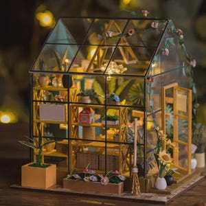 May include: A miniature greenhouse model with a glass roof and walls, featuring interior shelves filled with tiny plants, pots, and decorative items. The structure is framed in black and yellow, with a small door and a broom. The scene is illuminated with soft lights.