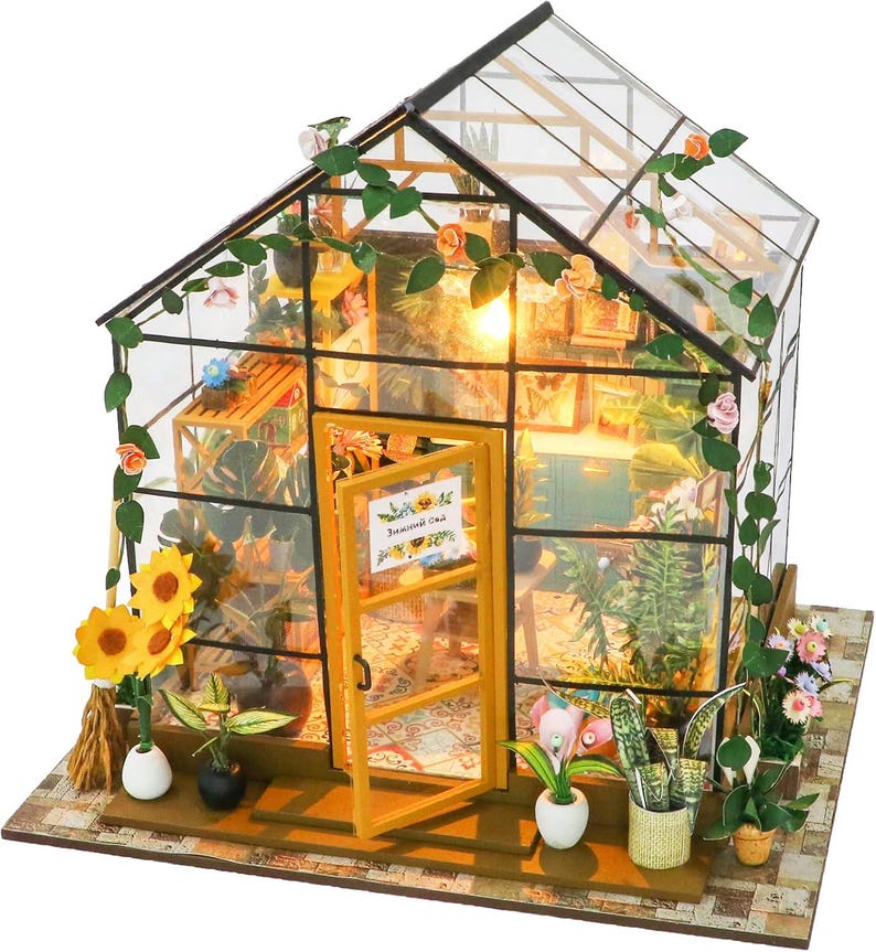 May include: A miniature greenhouse model with a glass roof and walls, featuring a wooden frame and door. The interior is filled with miniature plants, flowers, and decorative items. The exterior is adorned with artificial vines and flowers. The door has a sign.
